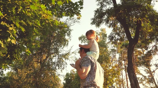 Babam parkta sevgili çocuğunun omuzlarını taşır. Baba, kızı ile birlikte ağaçların altında yürüyor. Ebeveynleri olan çocuk izin gününde yürür. Mutlu aile parkta dinleniyor.. — Stok fotoğraf