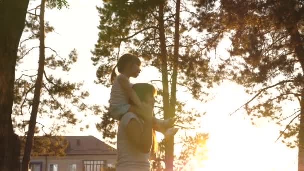 Mamma går med sin dotter på axlarna under träden. Mamma bär på sina axlar sitt älskade barn, i parken. barn med föräldrar går på lediga dagen. Lycklig familj är avkopplande i parken. — Stockvideo