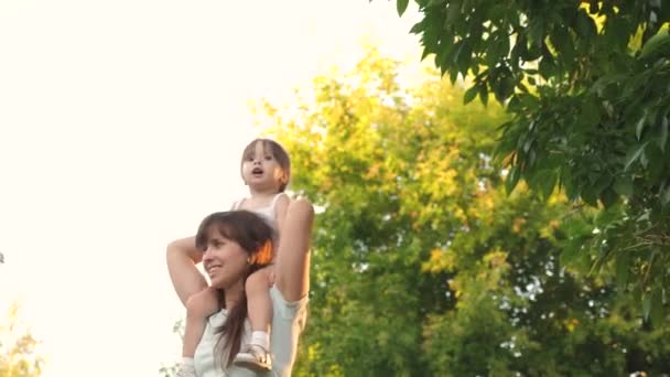 Moeder draagt haar geliefde kind op haar schouders, in het park. Mam loopt met haar dochter op haar schouders onder de bomen. kind met ouders loopt op de vrije dag. Gelukkige familie is ontspannen in het park. — Stockvideo