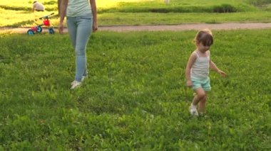 Küçük kız ve anne çimenlikte parkta yürürler. Çocuk yeşil çimlerin üzerinde yürüyor. Anne ve bebek çimlerde dinleniyor. Mutlu çocukluk kavramı. Mutlu ve çocuklu bir aile akşamları şehir dışına çıkar..