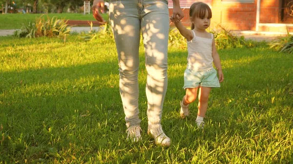 Little daughter and mother holding hands are walking in park on lawn. child walks on green grass. Mother and baby rest on lawn. concept of a happy childhood. A happy family is walking with a child. — Stock Photo, Image