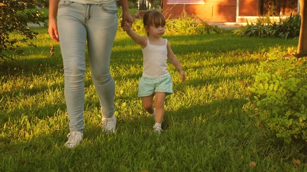 幼い娘と母親が芝生の上の公園を歩いています。子供は緑の草の上を歩く。母親と赤ん坊は芝生の上で休んでいる。幸せな子供時代の概念です。幸せな家族が子供と一緒に歩いている. — ストック写真