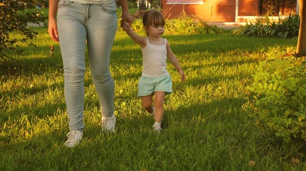 Little daughter and mother holding hands are walking in park on lawn. child walks on green grass. Mother and baby rest on lawn. concept of a happy childhood. A happy family is walking with a child. — Stock Photo, Image