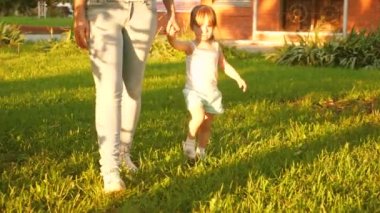 Küçük kız ve anne el ele tutuşuyorlar. Bahçede parkta yürüyorlar. Çocuk yeşil çimlerin üzerinde yürüyor. Anne ve bebek çimlerde dinleniyor. Mutlu çocukluk kavramı. Mutlu bir aile bir çocukla yürüyor..