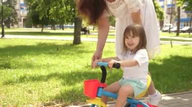 Çocuk bisiklet sürmeyi öğreniyor. Mutlu aile ve çocukluk kavramı. Ebeveynler ve küçük kızları parkta yürürler. Mutlu anne küçük kızına bisiklet sürmeyi öğretiyor. Anne dışarıda çocukla oynuyor..