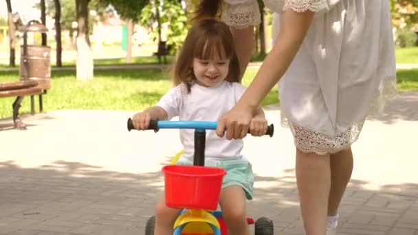 子供は自転車に乗ることを学びます。幸せな家族や子供時代の概念です。両親と娘は公園を歩いています。幸せな母親は自転車に乗るために娘を教えています。母親は屋外で子供と遊んでいます. — ストック動画