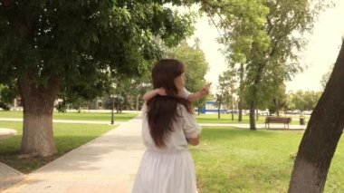 Güzel anne açık havada parkta küçük bir kızı taşıyor. Güzel anne ve bebeği parkta oynuyorlar. Bebek ve mutlu annesine sarılıyor. Anneler Günün kutlu olsun. Anne ve bebek. 4K