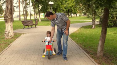 Babası çocuğuyla dışarıda oynuyor. Çocuk bisiklet sürmeyi öğreniyor. Mutlu aile ve çocukluk kavramı. Ebeveynler ve küçük kızları parkta yürürler. Mutlu baba küçük kıza bisiklet sürmeyi öğretiyor..