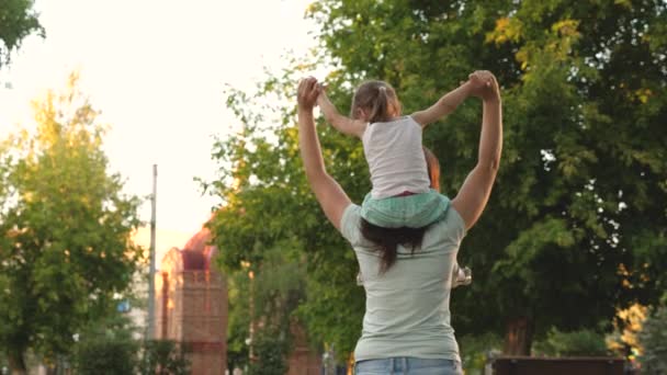 Мама ходит с дочерью на плечах под деревьями. Мать носит на плечах своего любимого ребенка, в парке. ребенок с родителями гуляет в выходной. Счастливая семья отдыхает в парке . — стоковое видео