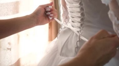 The bride dresses a white wedding dress. Bridesmaids tie the ribbons in the back of the brides dress. girl dresses a beautiful dress.