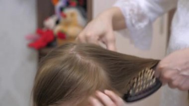 Kız için güzel saç stili kavramı. Mutlu bir aile. Anne, küçük kızının tomurcuğunu örüyor. Anne saçlarını bir tarakla tarıyor..