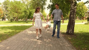 Mutlu çocuk babayla oynar ve anne ellerini tutar ve zıplar. Küçük kız parkta anne ve babasının elini tutarak zıplıyor. Aile kavramı. Doğada küçük bir çocukla yürümek.