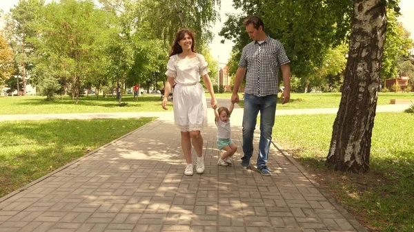 Criança feliz brinca com o pai e a mãe segura as mãos e pula. pequena filha pulando de mãos dadas de pai e mãe no parque. Conceito familiar. Caminhando com criança pequena na natureza . — Fotografia de Stock