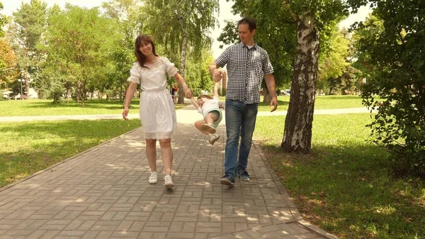 Szczęśliwe dziecko bawi się z tatą, a mama trzyma ich za ręce i skacze. Mała córeczka skacząca trzymając za ręce tatę i mamę w parku. Koncepcja rodziny. Spacerując z małym dzieckiem w naturze. — Zdjęcie stockowe