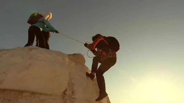 Équipe d'alpinistes grimpe une montagne sur une corde. femme libre aide un voyageur masculin à gravir une montagne. travail d'équipe de touristes. Voyage et aventure en montagne au coucher du soleil. les hommes d'affaires s'assurent mutuellement . — Photo