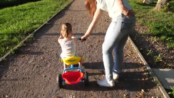 Mam leert dochter fietsen in het park. Een klein kind leert fietsen op een fiets. Moeder speelt met haar dochtertje. Het concept van een gelukkige jeugd. familievakantie in het weekend. — Stockvideo