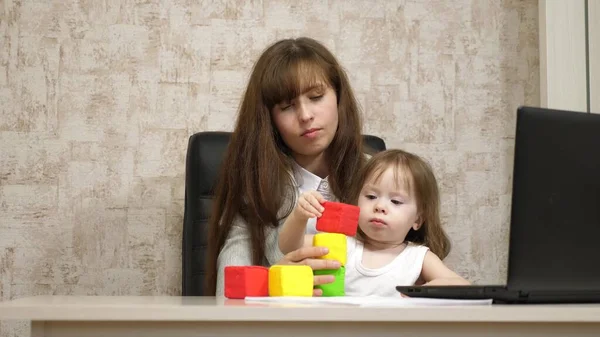 Trabajar en casa. en la habitación, mamá trabaja en la mesa con su hija pequeña en el ordenador. mujer de negocios que trabaja en la oficina en un portátil con un bebé en sus brazos. Trabajo independiente femenino . — Foto de Stock