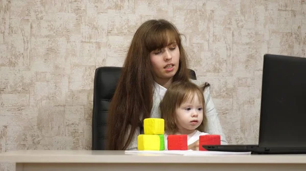 Trabalhar em casa. na sala, a mãe trabalha na mesa com a filha no computador. mulher de negócios trabalhando no escritório em um laptop com um bebê em seus braços. Trabalho feminino freelance . — Fotografia de Stock