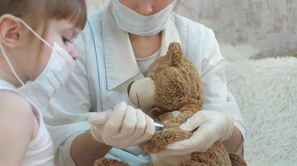 Cute girl makes an injection to toy bear. baby plays in a medical mask. child plays in hospital. game pretends to be doctor, nurse, veterinarian, heals a patient with a vaccine. danger of coronavirus — Stock Photo, Image