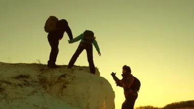 Bir grup dağcı birbirine yardım eli uzatarak bir dağa tırmanıyor. Özgür kadın gezgin dağa tırmanıyor. Turistlerin takım çalışması. Gün batımında dağlarda seyahat ve macera.