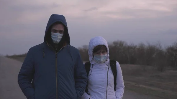 Woman and man escaping leave city with a backpack in protective medical mask and hood are walking outside city along road. Protection against viruses and bacteria. Security Concept, Coronavirus N1H1 — Stock Photo, Image