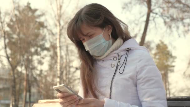 Молода жінка в медичній захисній масці сидить з смартфоном на вулиці міста в Європі. Захист коронавірусу. Туристи на вулиці носять захисну маску від вірусів. Здоров "я і безпека — стокове відео