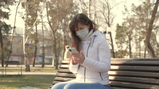 コロナウイルス対策。医療保護マスクの若い女性がヨーロッパの街の通りにスマートフォンを持って座っています。ストリートの観光客はウイルスから保護マスクを着用する。健康と安全を — ストック動画