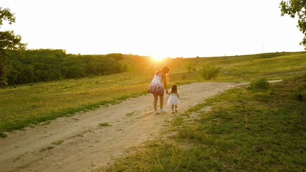Szczęśliwa rodzinna koncepcja. Mama i dziecko trzymają się za ręce i chodzą wieczorem po parku w promieniach pięknego zachodu słońca. Mała córeczka w białej sukience spaceruje z matką po drodze. szczęśliwa rodzina podróżuje — Zdjęcie stockowe