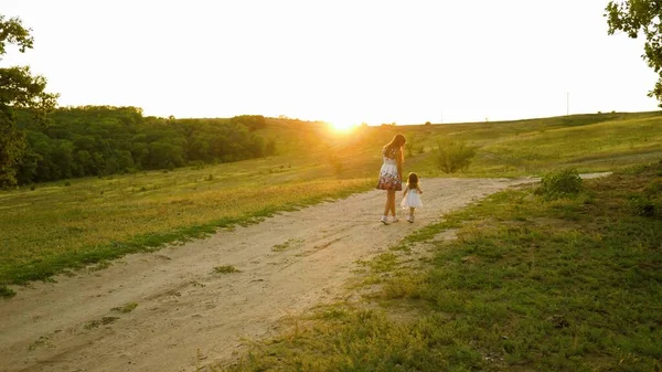 Concepto familiar feliz. Mamá y el bebé se toman de la mano y caminan por la noche en el parque en los rayos de una hermosa puesta de sol. Hijita con un vestido blanco camina con su madre en la carretera. familia feliz viajes — Foto de Stock