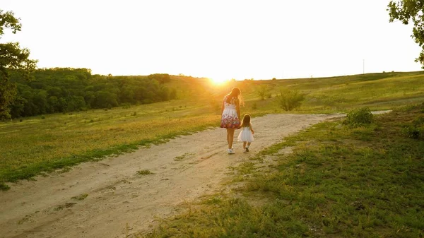 Szczęśliwa rodzinna koncepcja. Mama i dziecko trzymają się za ręce i chodzą wieczorem po parku w promieniach pięknego zachodu słońca. Mała córeczka w białej sukience spaceruje z matką po drodze. szczęśliwa rodzina podróżuje — Zdjęcie stockowe