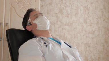 Klinikteki koruyucu maskeli ve gözlüklü yorgun doktor dinleniyor. Erkek doktor hastanede bir sandalyede uyuyakalmış. Sıhhiyeci gözlerini yorgunluktan ve fazla çalışmaktan kapattı.