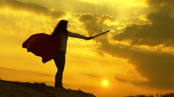 Mulher livre a fazer de super-herói. super mulher com uma espada na mão e em um manto vermelho fica em uma montanha na luz do pôr do sol. menina joga romanlenin em 9 raios brilhantes do sol contra o céu — Fotografia de Stock