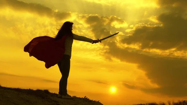 free woman playing a superhero. super woman with a sword in his hand and in a red cloak stands on a mountain in the sunset light. girl plays romanlenin in 9 bright rays of the sun against the sky