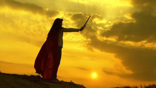 Wolna kobieta grająca superbohatera. super kobieta z mieczem w ręku i w czerwonym płaszczu stoi na górze w świetle zachodu słońca. dziewczyna gra romanlenin w 9 jasne promienie słońca przed niebem — Zdjęcie stockowe