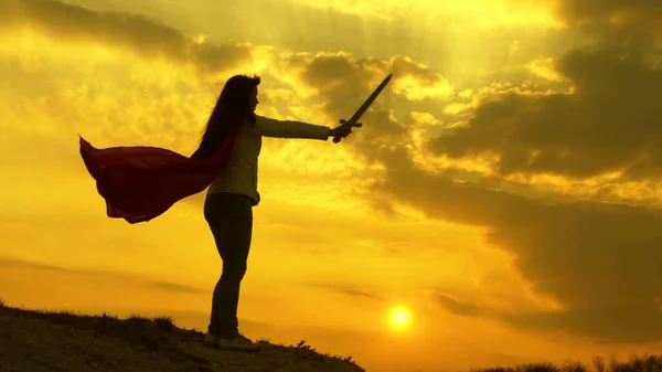 Femme libre jouant un super-héros. super femme avec une épée à la main et dans un manteau rouge se tient sur une montagne dans la lumière du coucher du soleil. fille joue romanlenin dans 9 rayons lumineux du soleil contre le ciel — Photo