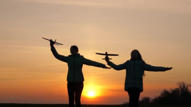 Silhuett av barn som leker på plan. Drömmar om att flyga. Glad barndomsidé. Två flickor leker med ett leksaksplan vid solnedgången. Barn på bakgrunden av solen med ett flygplan i handen. — Stockvideo