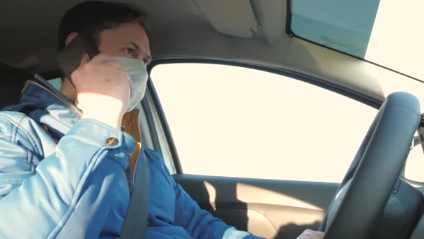 Gratis man met een mobiele telefoon reist met een masker in de auto. bescherming tegen virussen en bacteriën. De jongen rijdt en praat op Android. Gezonde jonge man in beschermende steriele medische masker rijdt een auto. — Stockvideo