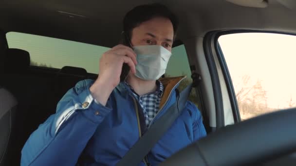 男の子はアンドロイドを運転して話している。保護滅菌医療マスクの健康な若い男は車を運転します。携帯電話を持つ無料の男は車の中でマスクで旅します。ウイルスや細菌からの保護. — ストック動画