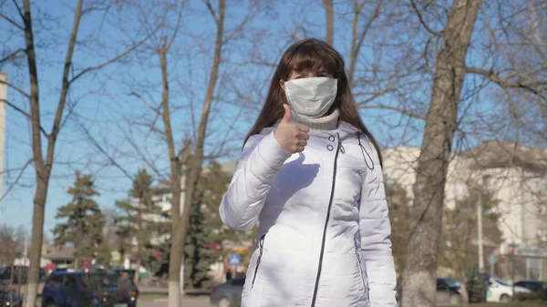 健康的女孩穿着防护的医疗面具在街上，显示喜欢。自由的女人戴着医疗面罩，带着病毒，上课。大流行性大肠癌的健康和安全概念 — 图库照片