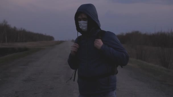 Omul liber care scapă din oraș cu un rucsac în mască medicală de protecție și capotă se plimbă în afara orașului de-a lungul drumului. om sănătos Protecție împotriva virușilor și bacteriilor. Security Concept — Videoclip de stoc