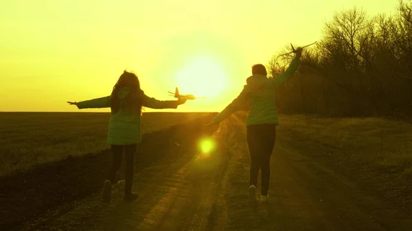 Due ragazze giocano con un aeroplano giocattolo al tramonto. sano I bambini corrono sulla strada con un aereo in mano. Silhouette di bambini gratis che giocano aereo. Sogni di volare. concetto di infanzia felice . — Foto Stock