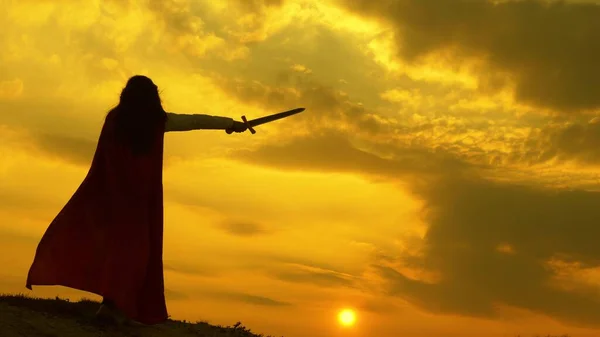 Vrije vrouwelijke ridder. super vrouw met een zwaard in zijn hand en in een rode mantel staat op de berg bij zonsondergang licht. vrije vrouw die superheld speelt. meisje speelt roman lenin in heldere stralen van zon tegen hemel — Stockfoto