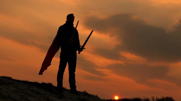 無料の男性騎士は手を伸ばして剣で祈る。剣を手にしたスーパーマンと赤いマントを着た男が夕日に照らされて山の上に立っている。フリーマンはスーパーヒーローだ。ローマ軍団のゲーム. — ストック写真
