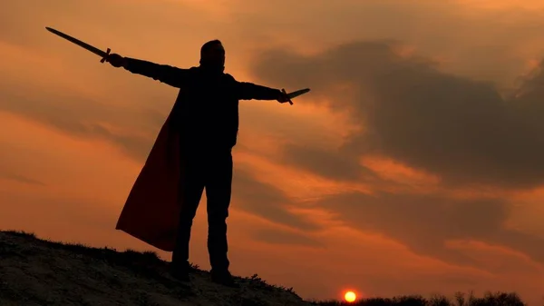 Szabad férfi lovag imádkozik karddal kinyújtott kézzel. Superman karddal a kezében és vörös köpenyben áll a hegyen napnyugtakor. A szabad ember szuperhőst játszik. játék római légió. — Stock Fotó