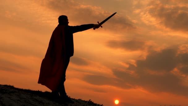 Özgür erkek şövalye. Elinde kılıçla ve kırmızı pelerinli Süpermen gün batımında bir dağın tepesinde duruyor. Özgür adam süper kahramanı oynuyor. Roma lejyonunun parlak güneş ışınlarıyla gökyüzüne karşı oyunu — Stok video