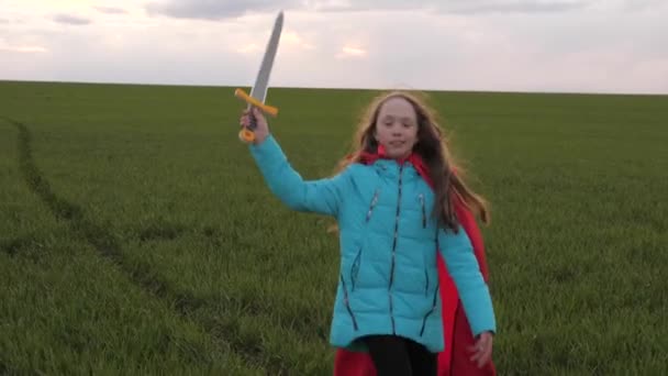 孩子们玩骑士游戏。穿着红色斗篷的女孩拿着剑穿过田野，扮演中世纪的骑士。孩子们用玩具剑战斗。快乐童年的概念。扮演超级英雄的小女孩 — 图库视频影像