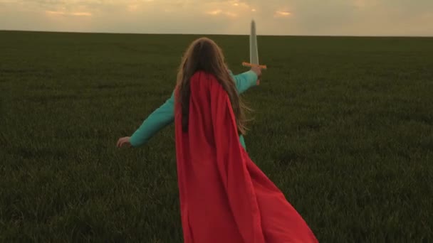 Leka riddare. flicka i en röd mantel löper med svärd i handen över fältet spelar medeltida riddare. Barn slåss med ett leksakssvärd. Glad barndom. ung flicka spelar superhjältar. — Stockvideo