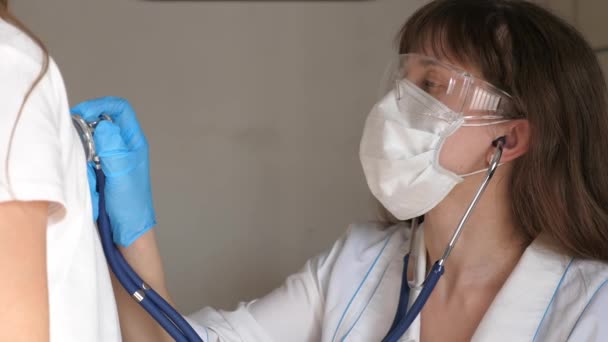 Arts met een beschermend masker en beschermende handschoenen onderzoekt de patiënt. close-up. Een vrouwelijke kinderarts met een stethoscoop luistert naar de longen en het hart van een meisje. geneeskundig concept. — Stockvideo