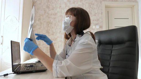 Μια γυναίκα γιατρός σε ένα γραφείο εξετάζει μια ακτινογραφία του ασθενούς στο νοσοκομείο. Ο γιατρός δουλεύει στο νοσοκομείο με λάπτοπ. Ιατρική περίθαλψη και υγειονομική περίθαλψη έννοια. πανδημία του κορονοϊού, πνευμονία COVID-19 — Φωτογραφία Αρχείου