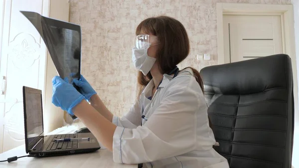 Μια γυναίκα γιατρός σε ένα γραφείο εξετάζει μια ακτινογραφία του ασθενούς στο νοσοκομείο. Ο γιατρός δουλεύει στο νοσοκομείο με λάπτοπ. Ιατρική περίθαλψη και υγειονομική περίθαλψη έννοια. πανδημία του κορονοϊού, πνευμονία COVID-19 — Φωτογραφία Αρχείου